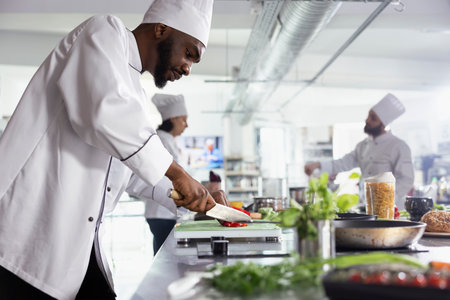 African american male chef slicing a red bell pepper on a cutting board with stainless steel tools in a restaurant kitchen. Man chopping delicious ingredients for a new gourmet dish.の写真素材