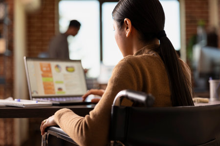 Rearview closeup of asian woman in wheelchair using laptop, concentrating on business analytics....