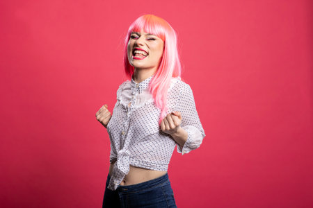 Joyful pink haired woman celebrating success, raising her fists in excitement and smiling brightly...