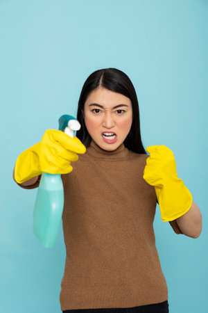 Stressed asian housekeeper wearing rubber gloves and holding cleaning spray stands in fight pose...