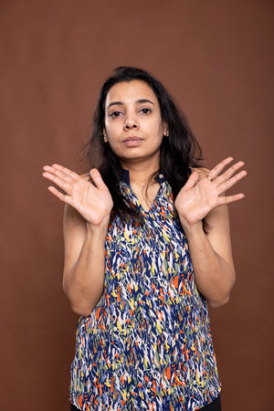 Focused indian woman standing with hands raised in rejection, firmly expressing disagreement. Her serious expression and confident posture convey assertive decision making in brown background studio.の写真素材