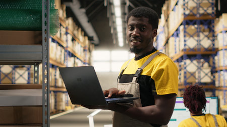 Black male staff verifying inventory pallets in e-commerce logistics center. Accurate order processing on laptop and packaging for quick shipping in large scale warehouse setting. Camera B.の写真素材