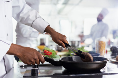 African american chef flipping the pork fillet in the pan using tongs, preparing the raw meat on the restaurant kitchen stove. Young male expert cooking steak in oil, food industry. Close up.の写真素材