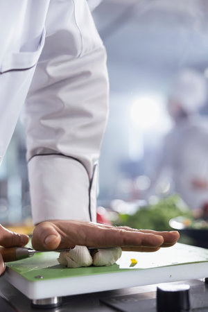 Culinary expert pressing the knife on garlic cloves at the kitchen counter, making crushed condiments and flavor on the cutting board. Young chef creates dish with tasty ingredients. Close up.の写真素材