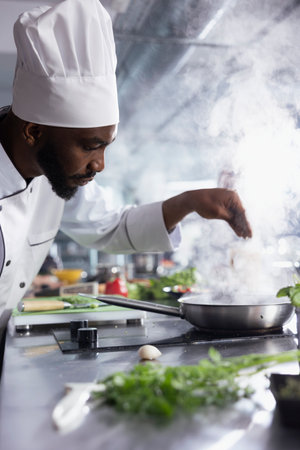 African american head chef sprinkles rosemary over a gourmet dish slow cooked on the stove, balanced gastronomy with fresh herbs. Kitchen counter filled with raw materials and fresh ingredients.の写真素材