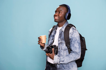 Male photographer poses with coffee cup and camera in hand, listening to music and carrying a backpack. Cheerful man wearing headphones stands against isolated backdrop, ready for photography session.の写真素材
