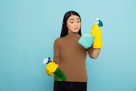 Asian janitor in yellow rubber gloves poses with spray bottles, standing against blue background. Female cleaner looking at liquid detergents in hand, preparing for efficient household sanitation.の写真素材