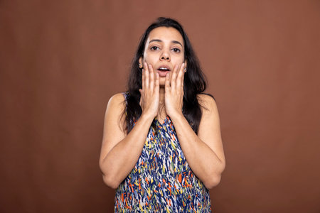 Beautiful indian lady expressing shock, hands on cheeks and mouth slightly open, reacting in amazement. Surprised woman with look of disbelief, staring directly at camera against isolated background.の写真素材