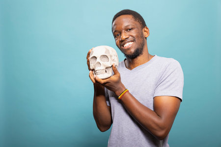 Cheerful black male person stands and grasps a skull close to him, posing for the camera in a...