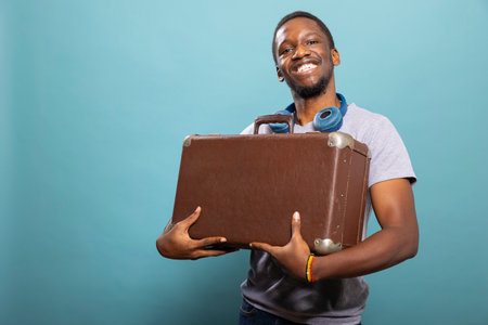 Black man cheerfully holding his brown suitcase, standing against blue background. African American tourist with headphones around neck, excited to travel as he carries his luggage close to his chest.の写真素材