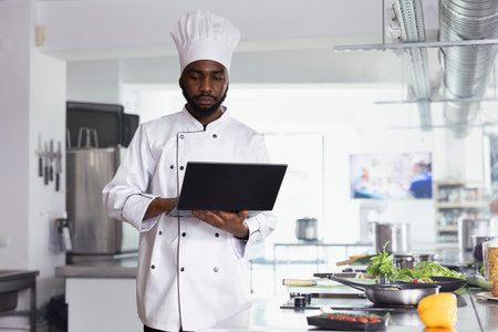 African american chef researching new cooking tips in restaurant kitchen uses a laptop while preparing fresh ingredients and plating food. High quality culinary arts and delicious dishes.の写真素材