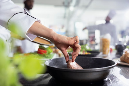 Culinary expert flipping the pork fillet in the pot with tongs and preparing the raw meat on the restaurant kitchen stove. Young professional cooking steak in oil, food industry. Close up.の写真素材