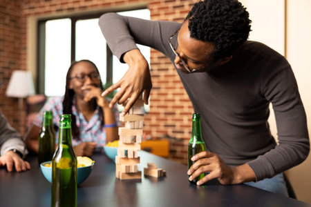 Young african american man stands, making bold move in jenga game while holding a drink in one hand. Focused and daring, black male individual pulls wooden block from the tower as friends watch.の写真素材