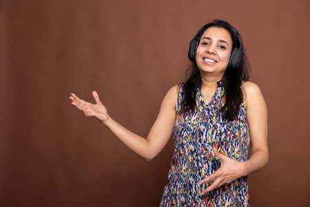 Playful female individual wearing wireless headphones, cheerfully dancing to music in brown background studio. Happy Indian woman enjoying the rhythm and miming an imaginary guitar.の写真素材