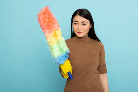 Smiling woman wearing yellow rubber glove and holding rainbow duster, determined to remove dust in...
