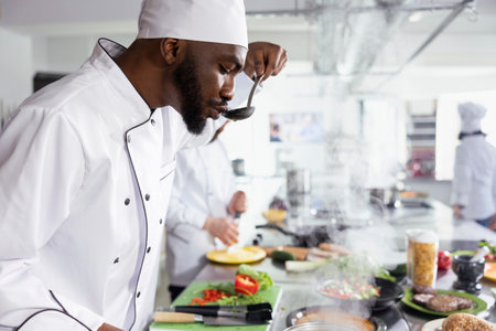 Black culinary expert tasting a pan of simmering sauce from the stovetop, making final adjustments to enhance rich and savory flavor. Chef life with delivering gourmet experiences.の写真素材