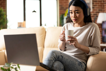 Self employed woman wearing wireless headphones, holding coffee mug while attentively watching online webinar on laptop. Remote female manager sitting in brick wall living room, focused and engaged.の写真素材
