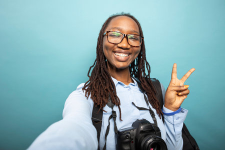 African american woman with DSLR around neck, showing peace sign to camera, filming content for...
