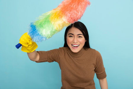 Young asian housekeeper with smile on face, excitedly holding feather duster, ready to tackle chores. Enthusiastic female cleaner prepared for professional household cleaning tasks.の写真素材