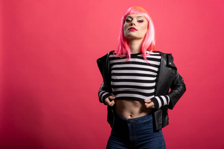 Stylish white woman with pink wig and striped crop top stands facing camera on isolated background. Confident female model in leather jacket shows bold attitude during fashion photoshoot in studio.の写真素材