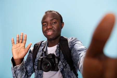 African American man with camera around his neck waving and greeting his online viewers, sharing...