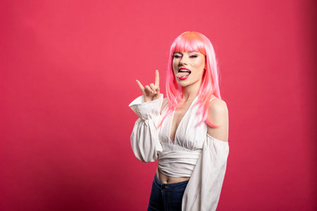 Fashion forward woman with pink hair sticks tongue out and flashes double horns hand sign, celebrating with energy and flair. Vibrant pink background complements her joyful and rebellious expression.の写真素材