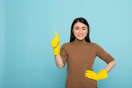 Cheerful female housekeeper poses proudly, showing thumbs up to express joy after completing chores. Happy asian cleaner in rubber gloves, hand on hip, confidently celebrating successful cleaning taskの写真素材