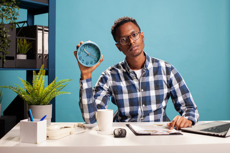 Portrait of african american freelancer sitting with a clipboard at desk and holding an alarm clock. Young business analyst handling paperwork and presenting the time with an analogue clock in hand.の写真素材