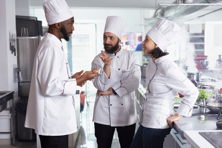 Culinary experts focus on cooking new dishes in a modern kitchen, wearing chef hats and uniforms. Diverse team sharing gourmet options at kitchen counter, preparing delicious meals.の写真素材