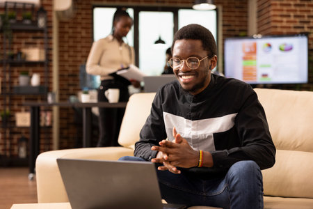 Black male employee seated on couch with a smile, watching live webinar on his laptop in brick wall...