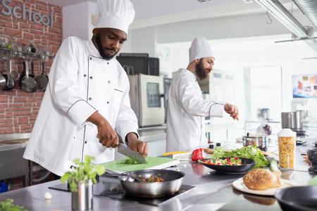 African american chef chopping herbs and spices on a cutting board, mincing raw ingredients for a fine dining dish. Young male cook using a big knife to slice fresh produce, gastronomy.の写真素材