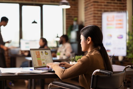 Female business professional in a wheelchair analyzing financial graphs on a laptop. Asian woman manager with a disability seated at office desk, leveraging technology to optimize company performance.の写真素材