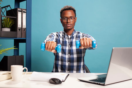 Male freelance sitting at the table and holding dumbbells towards the front, doing small exercises during work. African american man with glasses, using hand weights and looking at camera.の写真素材