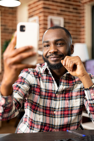 African american male manager holding mobile device, seated at desk, communicating remotely with coworker. Black businessman working from home, speaking with clients during video call on smartphone.の写真素材