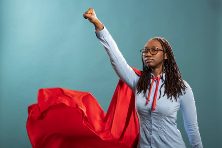 Confident woman stands in heroic stance, wearing red cape and blue shirt, exuding strength and empowerment. Black female model doing flying gesture, representing a superhero ready to save the day.の写真素材