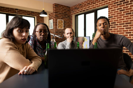 Friends unwind during evening downtime in indoor living space watching engaging story unfold with intensity and concentration. Young adults sitting together focused on laptop screen.の写真素材