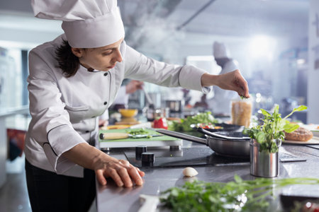 Female chef sprinkles fresh rosemary over a plated dish on the stove top, focusing on food plating and flavor. Preparing fresh ingredients, ensuring a blend of herbs in culinary workspace.の写真素材