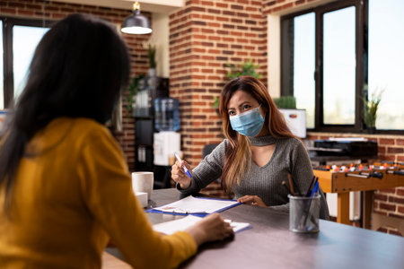 HR female representative with a pen in hand, seated at office desk conducting an interview meeting with potential job candidate. Asian businesswoman with face mask, having a discussion with employee.の写真素材