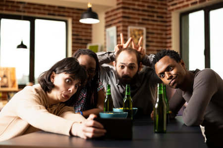 Diverse friends make goofy facial expressions while snapping selfies at table, as bearded man playfully shows bunny ears. Multiethnic group sit together, posing with smartphone, creating fun memories.の写真素材