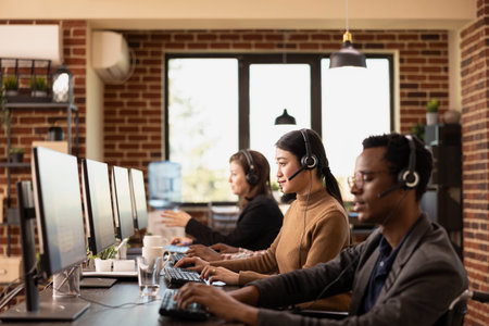 Selective focus on asian woman wearing a headset, seated among diverse coworkers and using computer in brick walled workspace. Multiethnic employees are working at their desks, speaking with clients.の写真素材