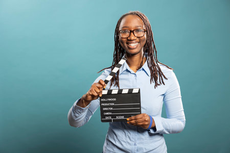 Cheerful female filmmaker looks at camera, holding a clapperboard, standing isolated over a blue...