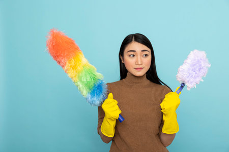 Young female janitor stands prepared with colorful cleaning tools during morning routine. Confident asian housekeeper ready for household chores, posing against isolated background.の写真素材
