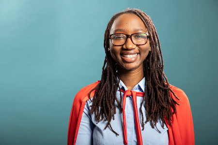 Joyful african american woman wearing red cape and glasses, expressing enthusiasm and confidence. She stands in blue background studio, embodying success, positivity and empowerment.の写真素材