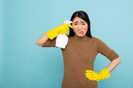 Overwhelmed asian housewife in yellow gloves holds cleaning spray bottle near forehead, expressing...