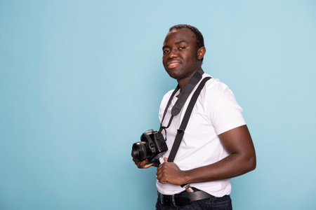 Smiling black male photographer holding his camera and gripping his suspenders as he stands in front of a blue background. Studio portrait perfect for creative, business, and lifestyle content.の写真素材