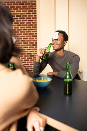 Smiling african american man sitting at table and holding beer bottle, casually hanging out with...