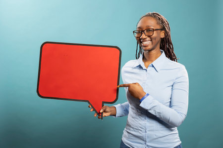 Joyful african american woman gesturing to blank speech bubble, expressing excitement over product. Smiling black female individual holds red cardboard sign, presenting copy space for advertisement.の写真素材