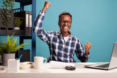 Excited manager cheering with his fist up after seeing positive results of his startup project. Black male entrepreneur celebrating after successfully sealing a business deal, arms raised in triumph.の写真素材