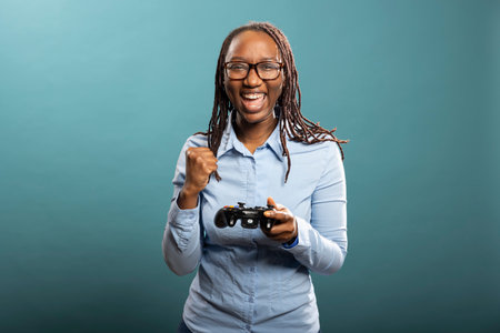 Cheerful black female individual with glasses, celebrating her video game win with fist raised....