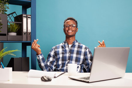 Smiling young man gesturing for cash by rubbing his fingers in a playful payday moment on a blue background. Cheerful black guy seated at his desk is expressing excitement over money rewards.の写真素材